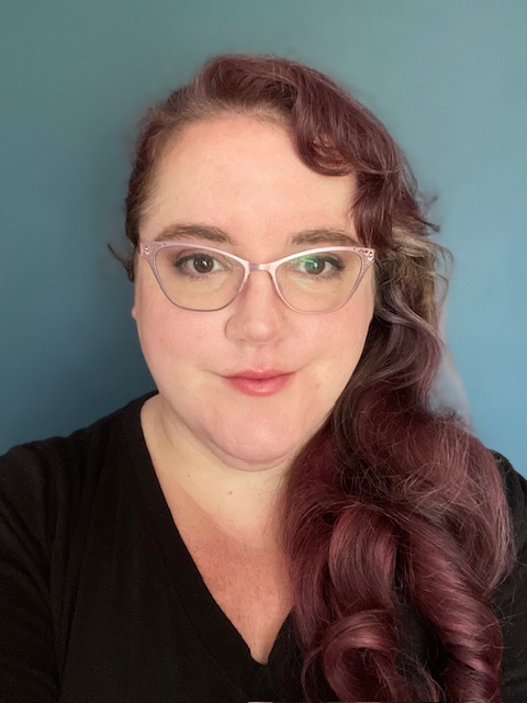 A woman with curly, shoulder-length hair tinted in shades of purple, wearing glasses and a black top, poses in front of a solid blue background.